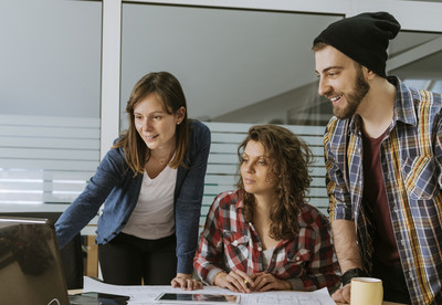 Foto: ein kleines Team arbeitet vor einem Computer an einem Projekt. Auf dem Tisch liegen Papierpl&auml;ne.