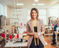 Foto: Eine Ausbilderin im Schneiderhandwerk in ihrem N&auml;hatelier. Sie schaut l&auml;chelnd in die Kamera, hat ein Ma&szlig;band um den Hals gelegt und ein Nadelkissen am Arm. 