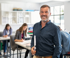 Foto: ein Lehrer an der Berufschule in seinem Klassenraum. Die Sch&uuml;ler/-innen im Hintergrund arbeiten an Aufgaben.