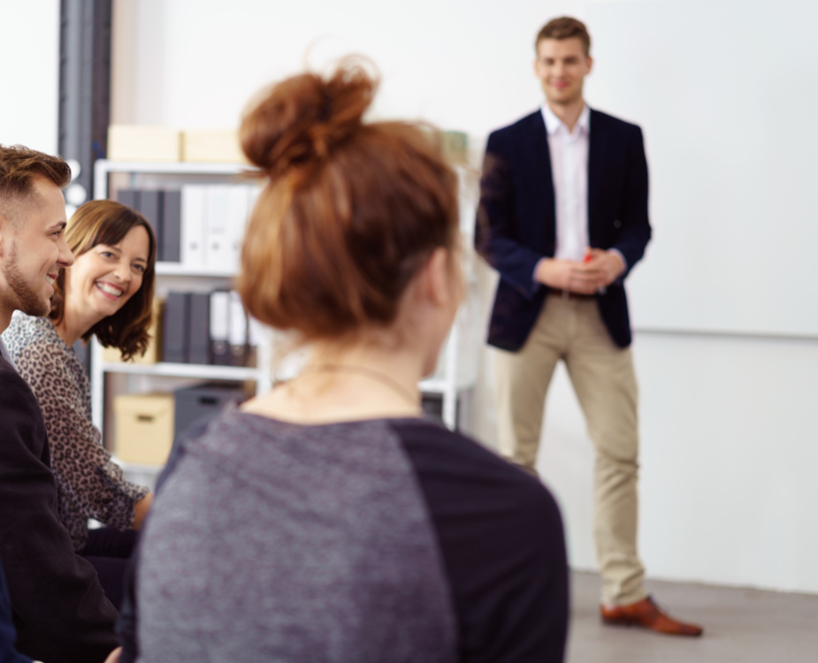 Foto: ein Auszubildender in Anzug h&auml;lt eine Pr&auml;sentation &uuml;ber seinen Auslandsaufenthalt vor Arbeitskolleginnen und -kollegen unterschiedlichen Alters.